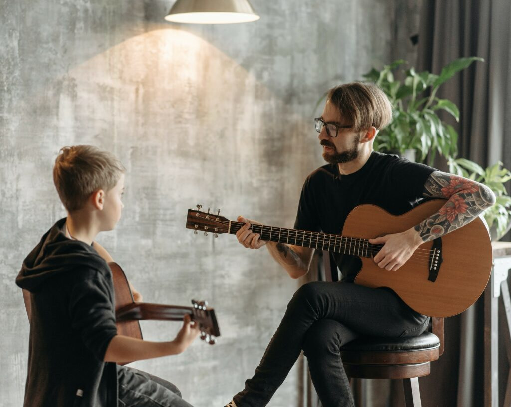 a man teacher with a guitar, sitting and showing a young boy with a guitar how to play