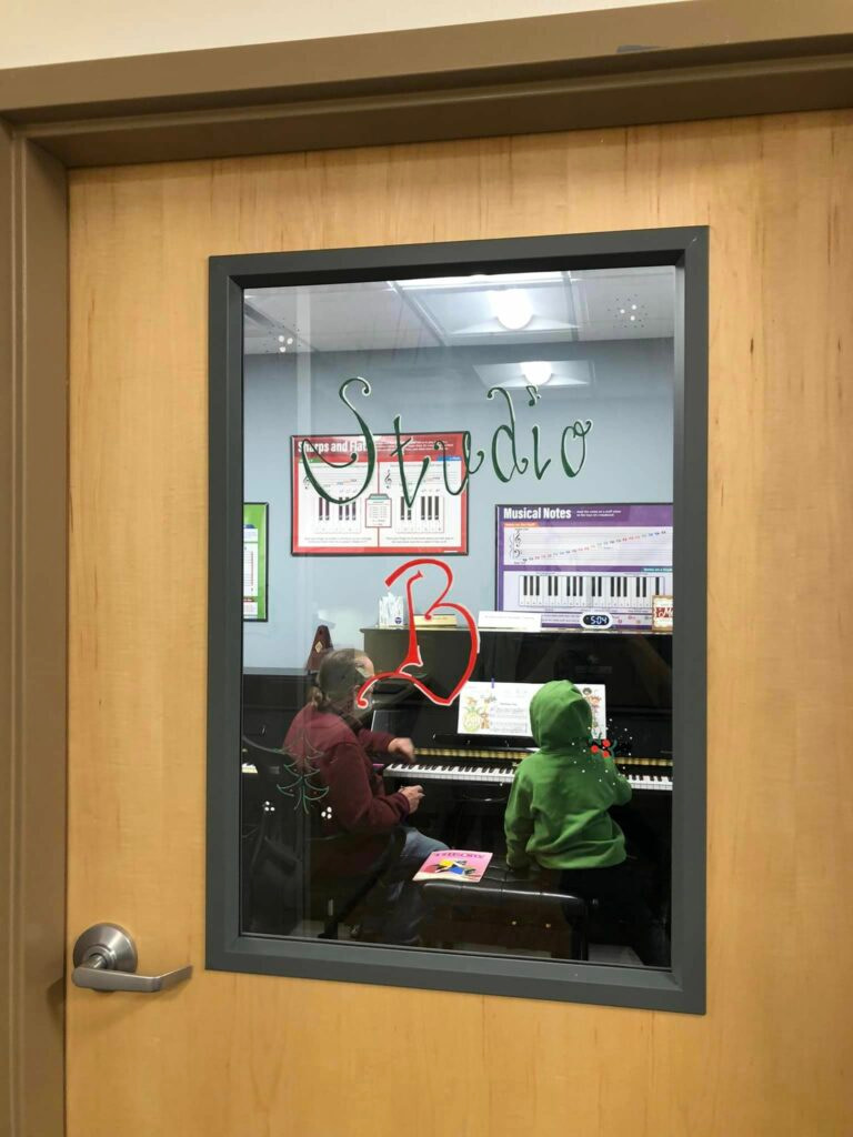 looking into the window of a practice room. A teacher and a young child with a green coat are at a piano.