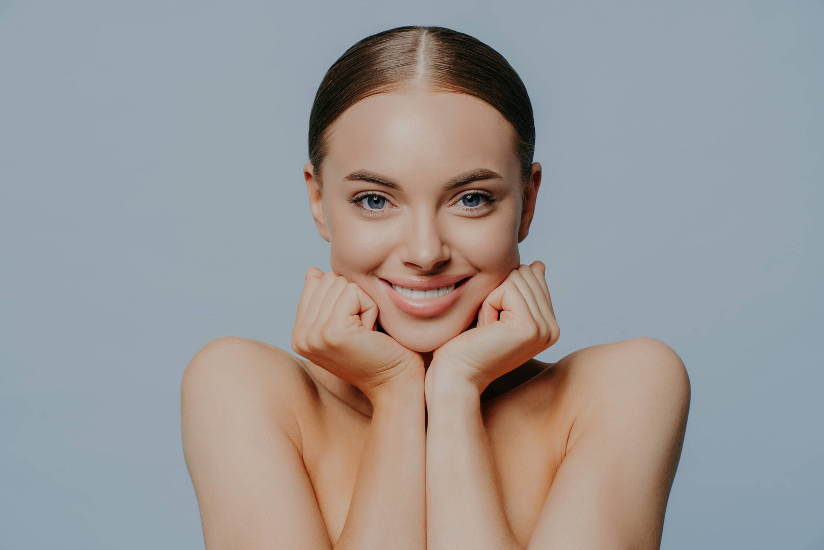 Women, Skin Care And Tenderness Concept. Young Female Takes Care Of Skin And Body Health, Keeps Hands Under Chin And Smiles Gently, Wears Makeup, Has Combed Hair, Delighted With Skin Care Product