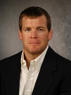 Tom Brands wearing a suit and tie smiling at the camera