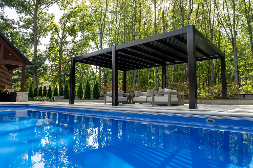 A large black pergola next to a pool on a backyard patio lined with tall trees.