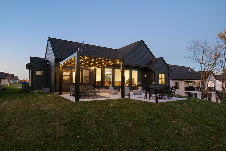 Black modern freestanding pergola over a patio is lit up at night by string lights