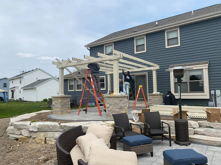 a landscaping crew is building a tan heartland pergola on top of stone pillars on an outdoor patio