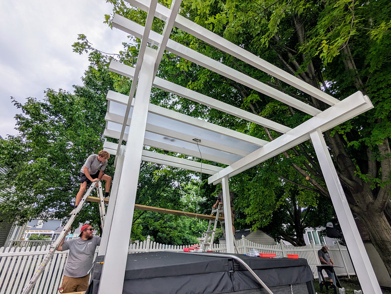 two men are building a white heartland pergola covered pergola over a hot tub.