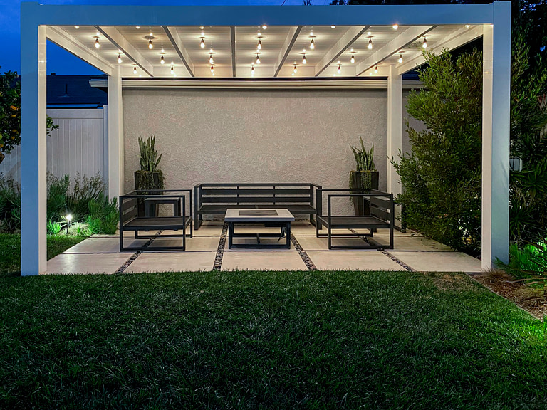 white modern pergola over a patio with string lights strung up under the pergola illuminating the space
