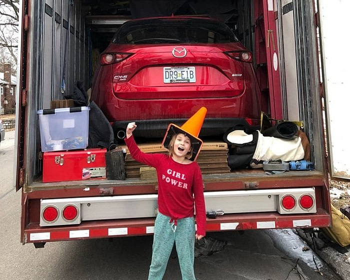 girl standing in front of moving truck with red car and other furniture inside